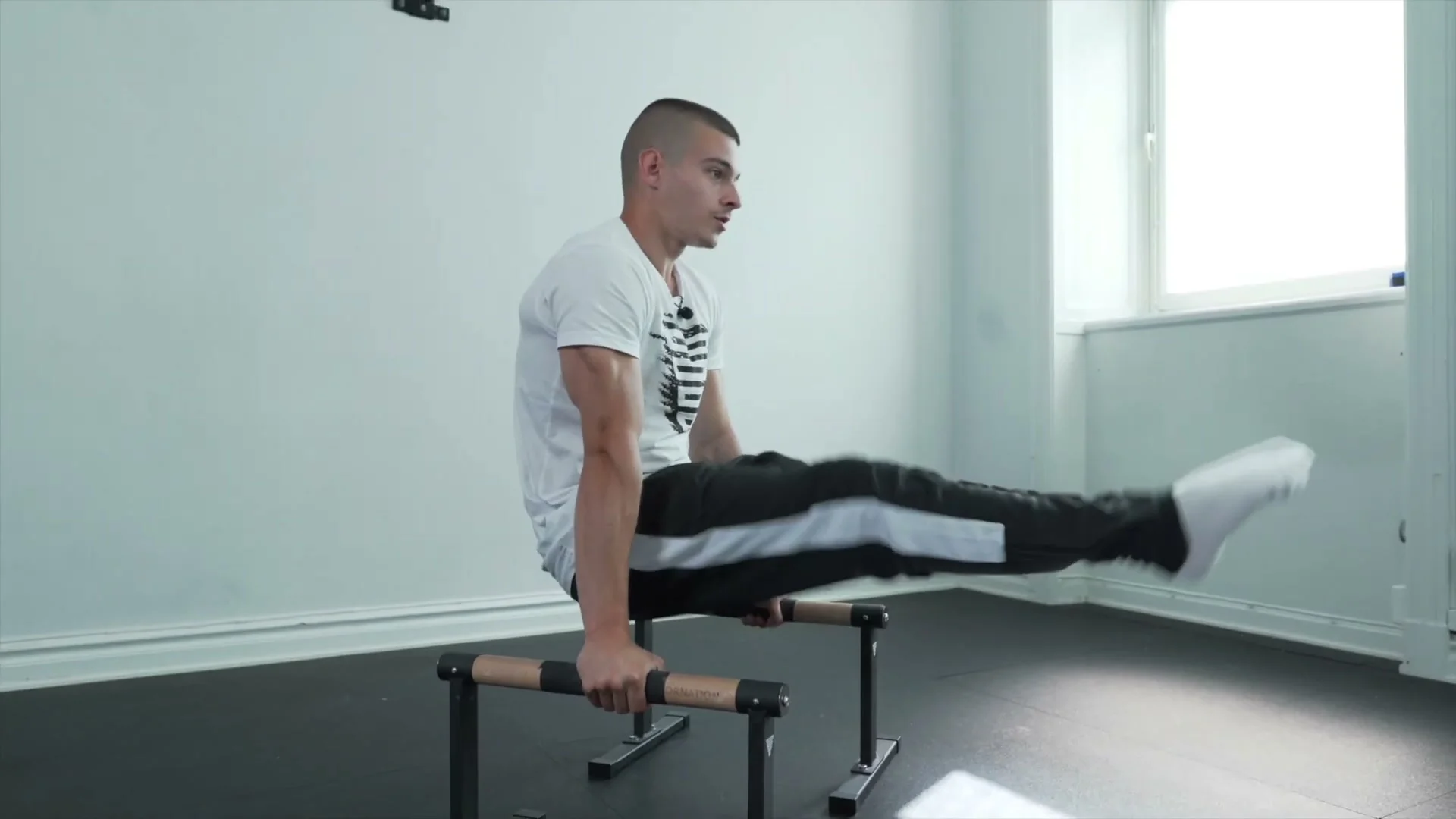 Daniel Hristov in the L-sit position on parallettes: body upright with legs extended horizontally forward, showing the starting position for the L-sit to straddle planche transition exercise